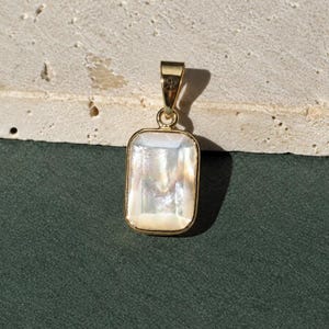 May include: A rectangular pendant featuring a polished, iridescent white mother-of-pearl inlay set within a smooth gold-toned metal frame. The pendant includes a small, integrated gold-toned bail at the top for attachment to a chain or cord.