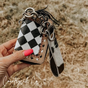 May include: A small, zippered wallet with a black and white chequered pattern. The wallet has a brown leather trim with a decorative edge and a wrist strap with a black and white cowhide pattern. The wallet is attached to a key ring with a silver clasp.
