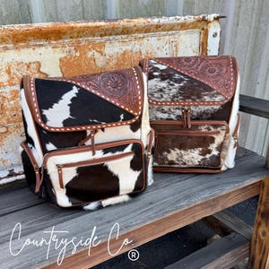 May include: Two cowhide backpacks with brown leather accents. One backpack has a black and white pattern, the other has a brown and white pattern. Both have a floral design on the flap and a front pocket with a zipper.