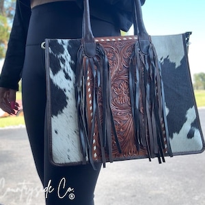 May include: A rectangular tote bag with a cowhide pattern on the sides and a brown, floral-tooled leather center. The bag has long brown handles and dark brown fringe. The text "Countryside Co." is visible at the bottom.