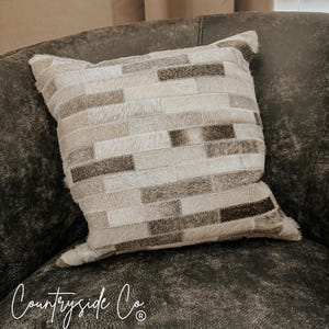 May include: A square decorative pillow with a patchwork design. The pillow features rectangular pieces of fur in shades of beige, gray, and brown. The pillow is resting on a dark brown leather couch. The text "Countryside Co." is written in cursive at the bottom.