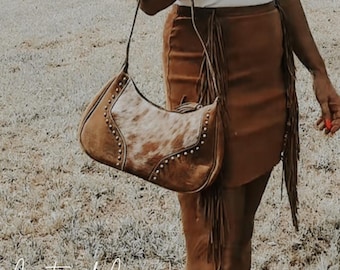 May include: A brown leather shoulder bag with a cowhide front and studded trim. The bag is worn with a brown suede skirt with fringe. The image includes the text "Countryside Co."