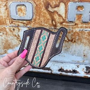 Puede incluir: Una funda de cuero marrón con un diseño decorativo turquesa y beige. La funda tiene un patrón floral y geométrico, con costuras blancas alrededor de los bordes. El logotipo de Countryside Co. es visible en la esquina inferior izquierda.