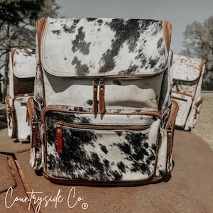 May include: A black and white cowhide backpack with brown leather accents and multiple pockets. The backpack has a zipper closure and the brand name "Countryside Co." is printed on the bottom.