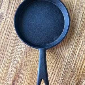 May include: A black cast iron skillet with a single handle. The skillet is small and round, with a smooth, black surface.