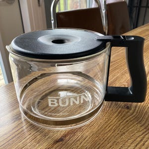 May include: A clear glass coffee pot with a black lid and handle. The pot has a gold band around the bottom and the word "BUNN" printed on the side.