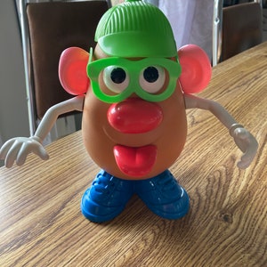 May include: A plastic Mr Potato Head toy with a green hat, green glasses, pink ears, a red nose, and a red tongue. The toy is standing on a wooden surface.