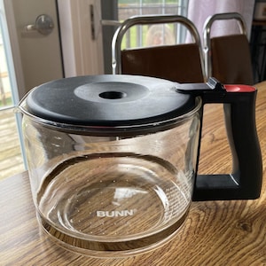 May include: A clear glass coffee pot with a black lid and handle. The pot has a black base and the word "BUNN" printed on the bottom.