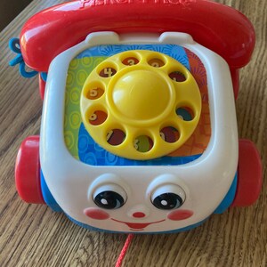 May include: A Fisher-Price toy telephone with a red receiver and wheels. The phone has a white face with a yellow rotary dial and a smiling face. It includes a pull string and is designed for young children.