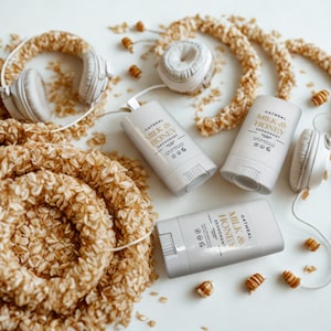 May include: Three white deodorant sticks labelled Oatmeal Milk & Honey, featuring a minimalist design with black text. The sticks are arranged alongside white over-ear headphones and circular wreaths covered in textured, light brown oat flakes.