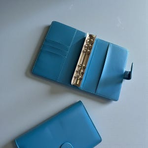 May include: A bright blue six-ring binder with an open interior featuring multiple card slots and a full-length pocket on the left side, and a matching closed blue binder positioned below it, both featuring a secure snap closure mechanism.