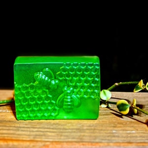 Pode incluir: Uma barra de sabão verde vibrante com um design de abelha e favo de mel. O sabão é retangular e apresenta entalhes detalhados de abelhas e um padrão de favo de mel. Um raminho de vegetação com folhas pequenas é colocado perto do sabão.