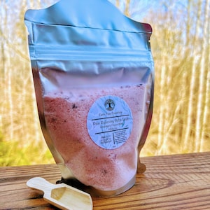 Puede incluir: Una bolsa de cierre hermético plateada llena de sales de baño rosas. La etiqueta dice "Earth Tone Essentials Pain Relieving Bath Soak Rosemary Peppermint Hibiscus Add 1-3 scoops to bathwater".