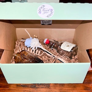 May include: A green gift box with a brown cardboard interior. The box contains a glass bottle with a cork stopper, a set of wooden diffuser sticks, and a decorative flower. The bottle has a label that reads "Honey & Vanilla".