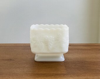 May include: White milk glass square bowl with a scalloped rim and a grape design. The bowl has a pedestal base.