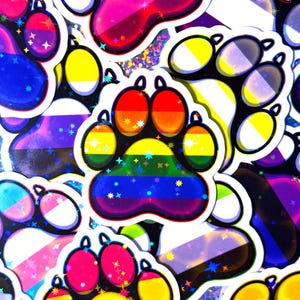 May include: A collection of colorful paw print stickers with rainbow, transgender, and other LGBTQ+ pride flag designs. The stickers have a holographic, glittery finish and are scattered across a black background.