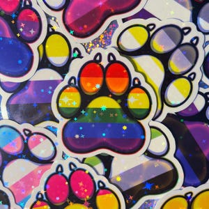 May include: A collection of colorful paw print stickers with various pride flag designs. The stickers feature rainbow, bisexual, transgender, non-binary, and other pride flag patterns. Each sticker has a holographic, glittery finish, creating a shimmering effect.