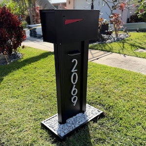 May include: A black mailbox with a red flag, mounted on a black post. The post features the address number "2069" in silver. The base is surrounded by white stones, set on a black frame, and is placed on a grassy lawn.