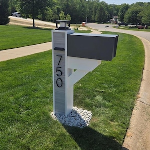 May include: A modern mailbox mounted on a white and gray wood-grain post featuring the house number 750 in black vertical lettering. The unit includes a dark gray rectangular mailbox, a black solar light fixture on top, and a white support bracket.