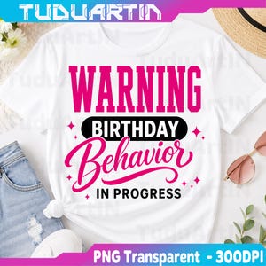 Puede incluir: Una camiseta blanca de manga corta con el texto en rosa intenso WARNING, un óvalo negro que contiene la palabra BIRTHDAY y el texto en cursiva rosa intenso Behavior IN PROGRESS, decorado con pequeñas estrellas rosas de cuatro puntas.