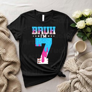 Puede incluir: Camiseta negra con el texto "BRUH I'M 7" en un dise&ntilde;o azul y rosa. El n&uacute;mero 7 tiene una combinaci&oacute;n de colores azul y rosa con un dise&ntilde;o de costura de b&eacute;isbol en la parte inferior. La camiseta es de estilo casual.
