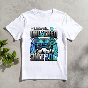 Pu&ograve; includere: T-shirt bianca con grafica colorata di un controller per videogiochi. Il design include il testo "LEVEL 9 UNLOCKED AWESOME SINCE 2017". Il controller &egrave; blu e verde.
