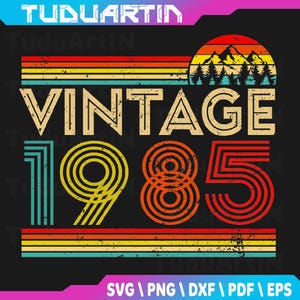 May include: A retro-style graphic design with the text "VINTAGE 1985" in beige, orange, and yellow. The design includes a sunset scene with mountains and trees, and horizontal stripes in various colors. The design is on a black background.