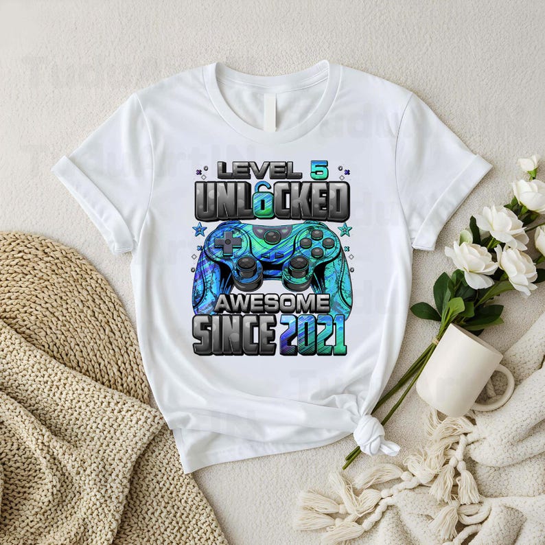 Pu&ograve; includere: T-shirt bianca con grafica colorata di un controller di videogiochi. Il design include il testo "LEVEL 5 UNLOCKED AWESOME SINCE 2021" in blu e verde, ideale per un compleanno da gamer.