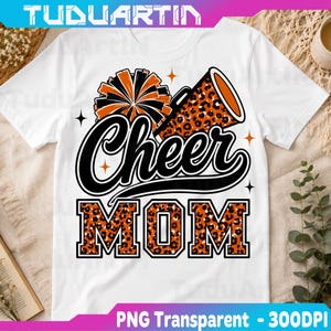 Op de afbeelding: Een wit T-shirt met korte mouwen en een grafisch ontwerp met een zwart-oranje pompon, een megafoon met luipaardprint en de tekst Cheer MOM in zwarte schrijf- en blokletters met een luipaardprintvulling, geaccentueerd door kleine oranje sterren.