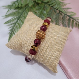 May include: A burgundy and gold beaded bracelet with a large gold rhinestone ball charm. The bracelet is displayed on a beige burlap pillow.