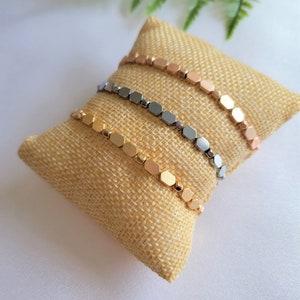 May include: Three bracelets with hexagonal beads in gold, silver, and rose gold colors. The bracelets are stacked on a beige burlap cushion.