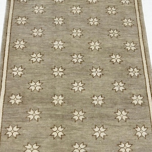 May include: A rectangular, beige rug with a repeating pattern of white floral designs. The rug has a cream-colored border along the edges. The floral pattern is evenly spaced across the rug's surface.