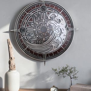 May include: A circular wall art piece featuring a layered compass design with a stylized wave and sun motif. The artwork incorporates metallic silver and dark brown tones, with the letters N and W clearly visible at the top and left cardinal points.