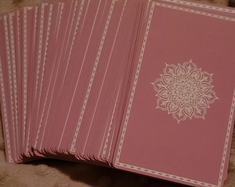 May include: A deck of tarot cards with a pink background and white border. The back of each card features a white mandala design. The cards are fanned out, showing the edges and the detailed design. The cards are a soft pink color.