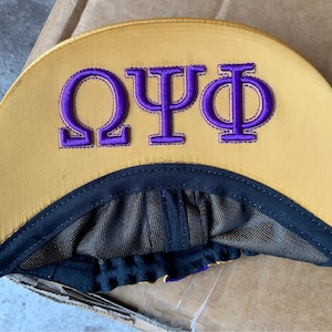 May include: A gold satin baseball cap with the Greek letters Omega, Psi, and Phi embroidered in purple. The cap has a black brim and a mesh lining. The hat is likely a fraternity or sorority item.