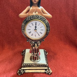May include: A decorative clock featuring a woman figure with arms raised, hands touching above the clock face. The clock has a gold-colored frame and Roman numerals. The base is square with a cream-colored top and gold accents. The letters "ΔΣΓ" are visible.