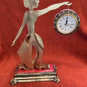 May include: A gold-toned art deco figurine of a woman holding a clock. The clock has a black and gold bezel with Roman numerals. The base is red with gold accents. The figurine is on a red surface.