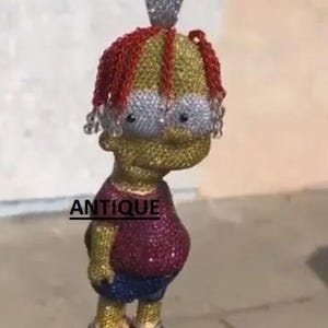 May include: A pendant necklace featuring a cartoon character design. The pendant is adorned with gemstones in various colors, including red, yellow, blue, and silver. The word "ANTIQUE" is printed on the pendant.
