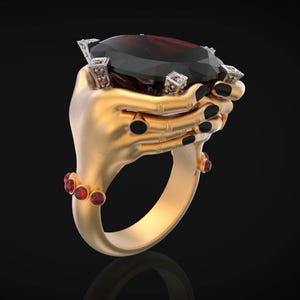 May include: A gold ring featuring a large, faceted, dark red gemstone held by two stylized hands. The hands have black nail polish and are adorned with small red gemstones. The ring is set against a black background.