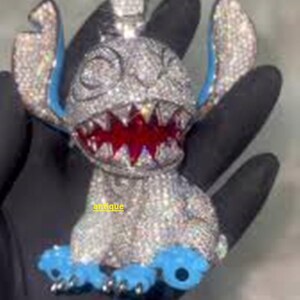 Puede incluir: Un colgante con incrustaciones de diamantes con forma de personaje de dibujos animados con orejas y pies azules y boca roja. El colgante se sostiene en una mano enguantada de negro. La palabra "antique" es visible.