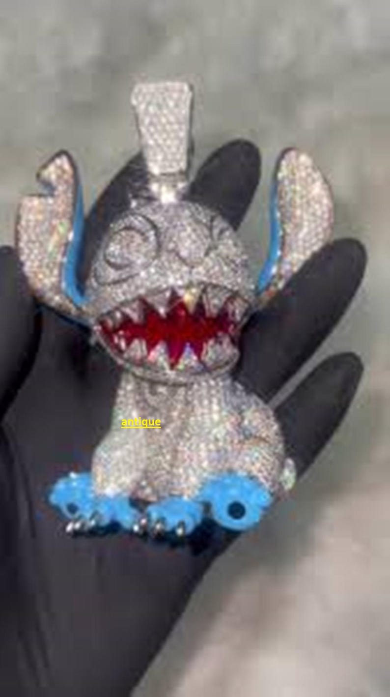 Puede incluir: Un colgante con incrustaciones de diamantes con forma de personaje de dibujos animados con orejas y pies azules y boca roja. El colgante se sostiene en una mano enguantada de negro. La palabra "antique" es visible.