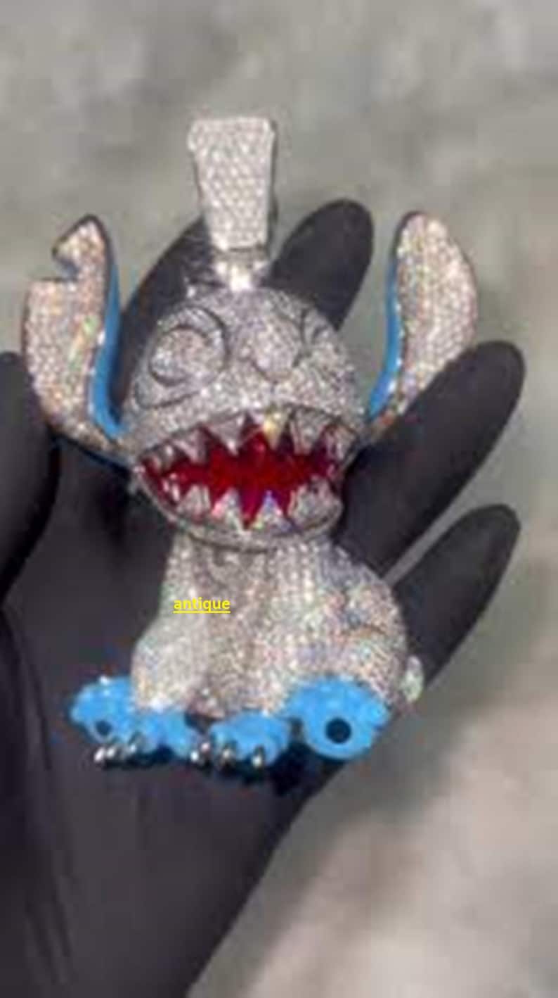 Puede incluir: Un colgante con incrustaciones de diamantes con forma de personaje de dibujos animados con orejas y pies azules y boca roja. El colgante se sostiene en una mano enguantada de negro. La palabra "antique" es visible.