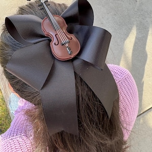 May include: A large black hair bow with a miniature brown violin attached. The bow is made of a wide, ribbed fabric. The violin has detailed strings and a small black bow. The hair accessory is worn on a person's head.