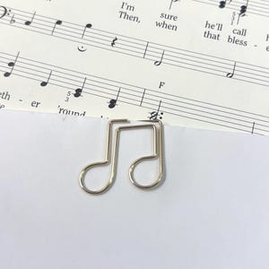 Peut inclure: Un trombone argenté en forme de note de musique est posé sur une partition. La partition comporte des notes de musique et les mots "nests? vine. I'm Then, sure when he'll call that bless".