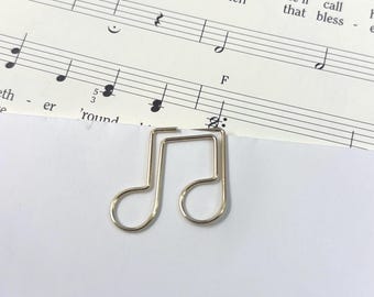 May include: A silver-colored music note paper clip rests on sheet music. The sheet music has musical notes and the words "nests? vine. I'm Then, sure when he'll call that bless". The paper clip is in the shape of a musical note.