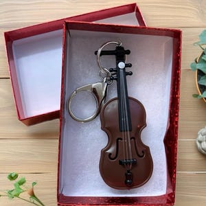 May include: A brown violin keychain with black strings and details, presented in a red gift box. The keychain is made of a shiny material and has a silver-colored keyring. The box is open, revealing a white interior.