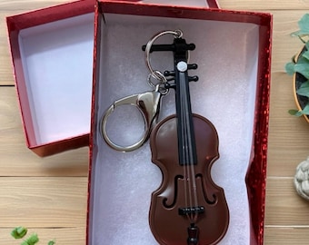 Puede incluir: Un llavero de violín marrón con cuerdas negras, presentado en una caja de regalo roja. El llavero es de material brillante y tiene un anillo plateado. La caja está abierta, revelando un interior blanco.