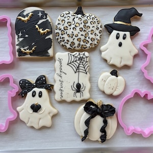 以下が含まれることがあります： ハロウィーンをテーマにしたクッキーとピンクのプラスチック製クッキー型のセット。クッキーのデザインには、金色のコウモリが描かれた黒い墓石、ヒョウ柄のパンプキン、魔女の帽子をかぶったゴースト、クモの巣と「spooky season」の文字、小さな白いパンプキン、黒いリボンをつけたゴースト、黒いリボンをつけた白いパンプキンが含まれています。