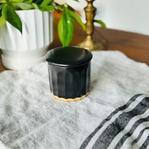 May include: A black ceramic jar with a gold rim and a matching lid. The jar is sitting on a white and black striped fabric.