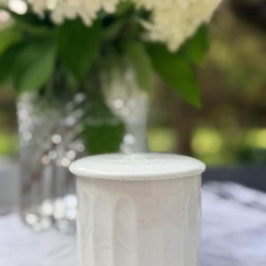 May include: A cylindrical white ceramic jar with a matching lid, featuring a subtle speckled texture and vertical fluted grooves along the exterior body. The container has a smooth, glossy finish and a clean, simple design suitable for storage.